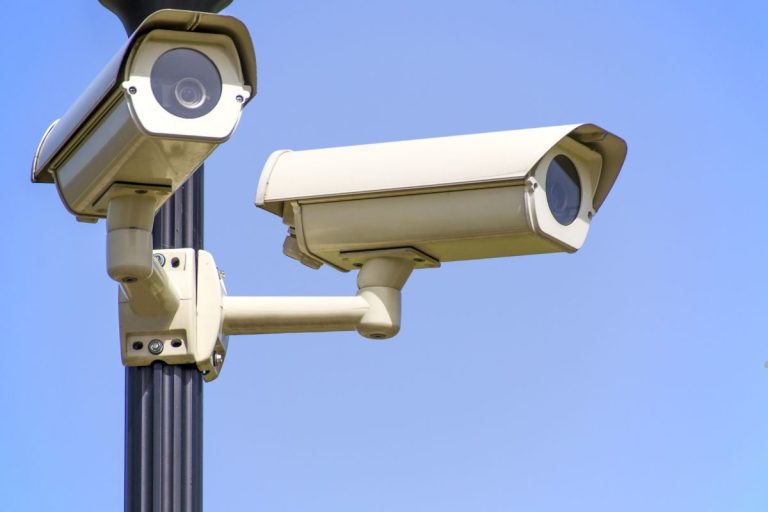 Two cream-colored security cameras mounted on a black pole against a clear blue sky, monitoring the surroundings.