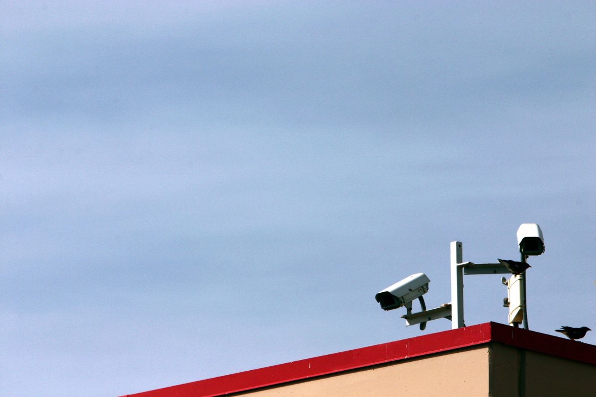 Two security cameras mounted on a building with a red roof, under a clear blue sky, and birds perched nearby.