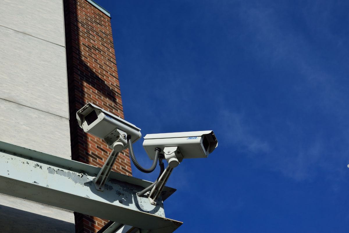 Two surveillance cameras mounted on a metal bracket, overlooking a brick building against a clear blue sky.